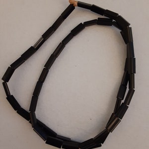 Könnte beinhalten: Eine schwarze Perlenkette aus rechteckigen Perlen. Die Kette ist auf einer dünnen Schnur aufgefädelt und hat einen einfachen Knoten am Ende.