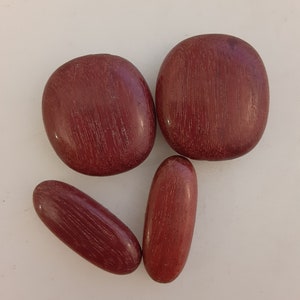 May include: Four smooth, rounded, red wooden stones. The stones are arranged in a loose pattern on a white surface.