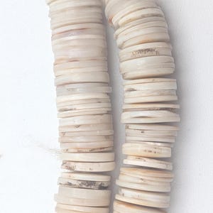 May include: Two strands of off-white, flat, circular beads are shown against a white background. The beads are strung together with a natural-colored cord. The beads have a slightly iridescent quality and vary in color from cream to light brown.