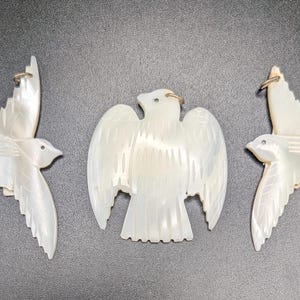 May include: Three white iridescent mother-of-pearl pendants carved into bird shapes. Two pendants depict birds in flight with wings extended, while the central pendant features a bird with wings spread wide. Each piece includes a small metal jump ring.