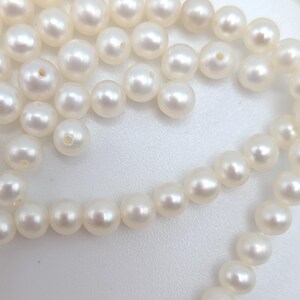 May include: A collection of round, off-white pearls arranged in a loose cluster. Each individual pearl features a smooth, lustrous surface and a small, centered drill hole, showcasing the natural spherical shape and consistent creamy color of the beads.