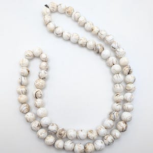 Puede incluir: Un collar compuesto por cuentas blancas y redondas con vetas marrones. Las cuentas están ensartadas, formando una doble hebra. El collar se muestra sobre un fondo blanco, resaltando los patrones y colores naturales de las cuentas.