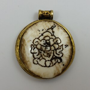 Puede incluir: Un colgante redondo de hueso blanco con un marco de metal dorado. El colgante presenta un diseño grabado detallado de una figura estilizada.