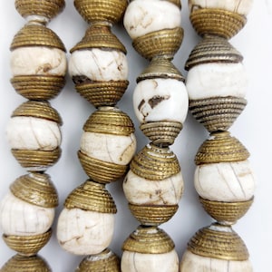 Puede incluir: Una hebra de cuentas blancas y doradas. Las cuentas son redondas y tienen una superficie texturizada. Las cuentas están enhebradas en un cordón y están dispuestas en una fila.