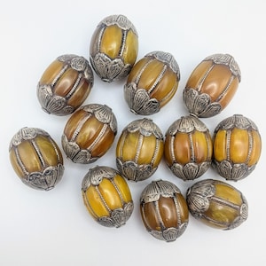 May include: A collection of oval-shaped beads with a silver-colored metal cap on each end. The beads have a warm, amber-like color with vertical grooves. The metal caps feature a floral design. The beads are arranged on a white surface.