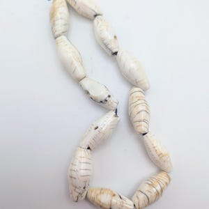 Puede incluir: Un collar compuesto por cuentas oblongas de color blanco roto con estrías marrones. Las cuentas están ensartadas en un hilo negro, creando un diseño único y naturalista. El collar se presenta sobre un fondo blanco liso.