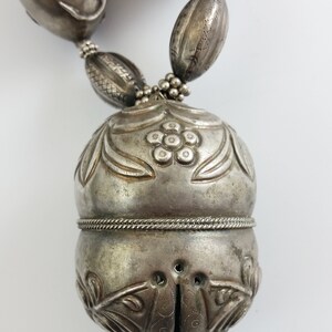 May include: A silver necklace with a large, ornate pendant. The pendant is shaped like a bell and has intricate floral designs. The necklace is made of silver beads and has a silver clasp.