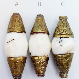 Puede incluir: Tres cuentas de concha blanca con tapas de metal dorado. Las tapas tienen diseños intrincados y están ligeramente desgastadas. Las cuentas son de forma ovalada y tienen una superficie lisa y pulida.