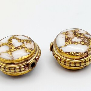 Puede incluir: Dos cuentas redondas de color dorado con diseños moteados en blanco y dorado. Las cuentas tienen un agujero en el centro para enhebrar.