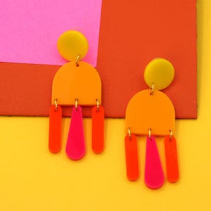 May include: A pair of vibrant, geometric earrings. Each earring features a yellow circle, an orange half-circle, and dangling shapes in orange and pink. The earrings are set against a colorful background of pink, orange, and yellow.