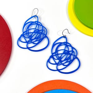 May include: A pair of abstract blue earrings with a tangled, looped design. The earrings are made of a solid material and hang from silver-coloured hooks. The background includes colourful circular shapes in red, orange, yellow, and green.