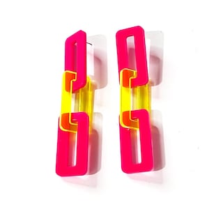 May include: A pair of vibrant, geometric earrings. Each earring features a chain-like design with bright pink rectangular links, interspersed with translucent yellow and orange links. The earrings are set against a white background, highlighting their bold colors and modern style.