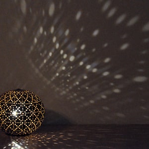 May include: A black metal lampshade with a geometric pattern casts a shadow of light on a wall. The lampshade is round and has a small light source inside.