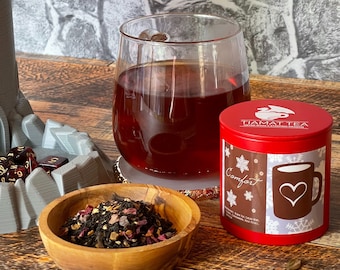 May include: A red metal tin of Comfort tea from Tiamat Tea. The tin features a snowflake design and the words "Comfort" and "Tiamat Tea" printed on it. A glass of red tea and a bowl of loose leaf tea are also in the image.