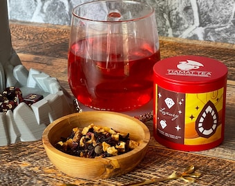 May include: A red tea tin with gold accents and the text "Tiamat Tea" and "Luxury" on the front. The tin is sitting on a wooden table next to a glass of red tea, a small wooden bowl filled with loose leaf tea, and a gold spoon.