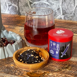 Puede incluir: Una lata de té roja con el texto "Tiamat Tea" y "Tradition" en ella. La lata está llena de té negro con aciano y té negro. También hay un vaso de té rojo y un cuenco de madera con té suelto en la imagen.
