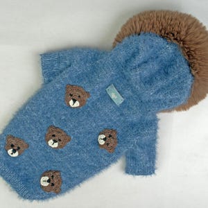 Puede incluir: Un suéter azul difuso para mascotas con capucha y borde de piel sintética marrón. El suéter está decorado con múltiples aplicaciones pequeñas de caras de oso tejidas a ganchillo en color marrón y tiene una pequeña etiqueta rectangular azul.
