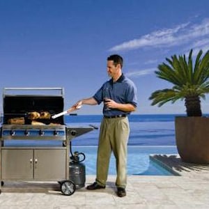 May include: A stainless steel gas grill with four burners and a side table. The grill is set up on a patio overlooking a pool and the ocean. A person is grilling food on the grill and another person is sitting on a chair nearby.