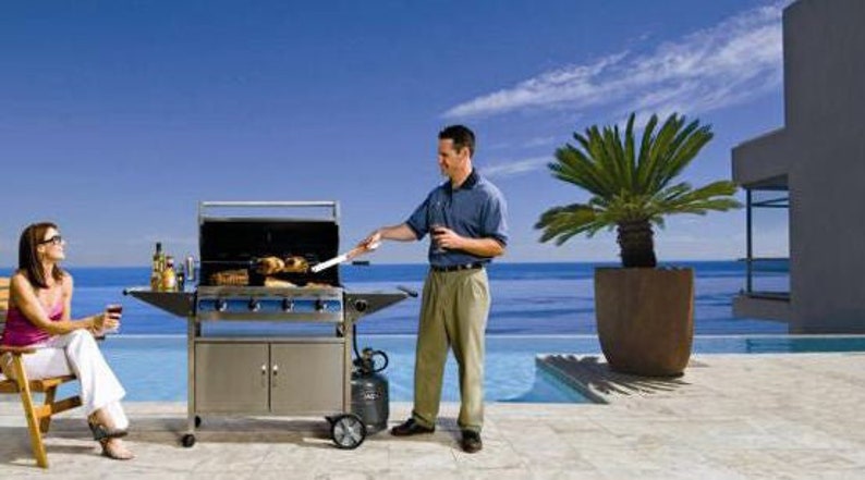 May include: A stainless steel gas grill with four burners and a side table. The grill is set up on a patio overlooking a pool and the ocean. A person is grilling food on the grill and another person is sitting on a chair nearby.