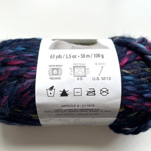 Puede incluir: Ovillo de lana azul oscuro con motas rosas, turquesas y amarillas. La etiqueta indica que es s&uacute;per voluminoso, 58 metros y 100 gramos. Adecuado para un ganchillo de 9 mm y agujas US 13.