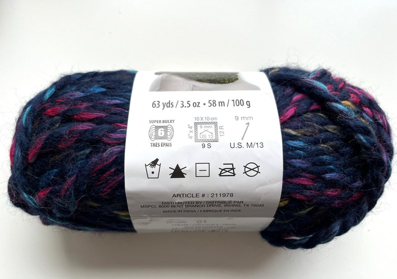 Puede incluir: Ovillo de lana azul oscuro con motas rosas, turquesas y amarillas. La etiqueta indica que es s&uacute;per voluminoso, 58 metros y 100 gramos. Adecuado para un ganchillo de 9 mm y agujas US 13.
