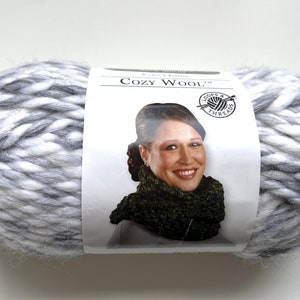 Puede incluir: Un gran ovillo de lana Cozy Wool en tonos blanco, gris y antracita. La lana es gruesa y voluminosa, ideal para tejer o hacer ganchillo. La etiqueta muestra a una mujer con un cuello tejido.