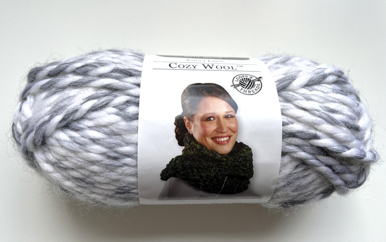 Puede incluir: Un gran ovillo de lana Cozy Wool en tonos blanco, gris y antracita. La lana es gruesa y voluminosa, ideal para tejer o hacer ganchillo. La etiqueta muestra a una mujer con un cuello tejido.