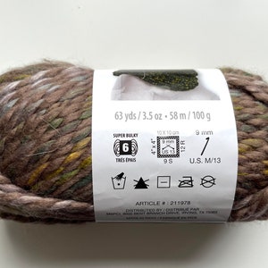 Puede incluir: Ovillo de lana s&uacute;per gruesa en tonos marrones, verdes y amarillos. La etiqueta indica 58 metros, 100 gramos y un tama&ntilde;o de aguja de 9 mm. Art&iacute;culo #211978.