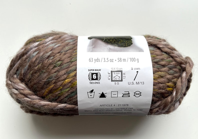 Puede incluir: Ovillo de lana s&uacute;per gruesa en tonos marrones, verdes y amarillos. La etiqueta indica 58 metros, 100 gramos y un tama&ntilde;o de aguja de 9 mm. Art&iacute;culo #211978.