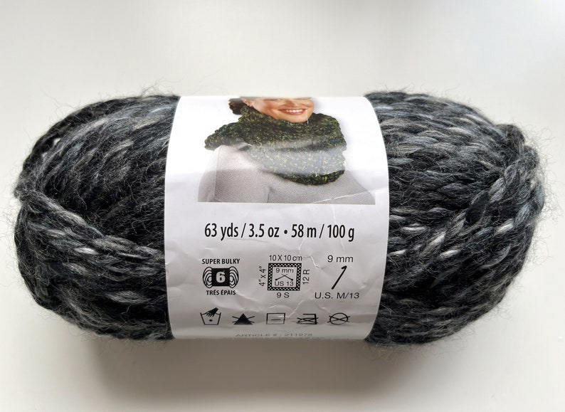 Puede incluir: Ovillo de lana s&uacute;per voluminosa en tonos grises y blancos. La etiqueta indica 58 metros, 100 gramos y recomendaciones de tama&ntilde;o de aguja. El grosor de la lana es 6.