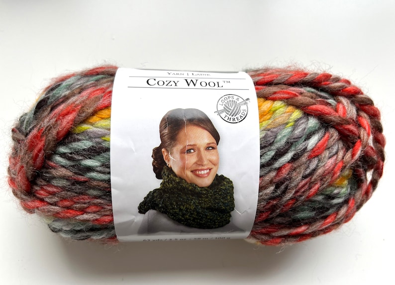 Puede incluir: Un gran ovillo de lana Cozy Wool en una mezcla de colores que incluye rojo, marr&oacute;n, amarillo y gris. La etiqueta muestra a una mujer con una bufanda verde. La lana pesa 180 g y mide 128 m.