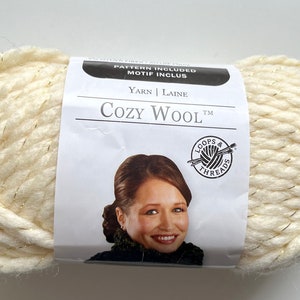 Puede incluir: Ovillo de lana Cozy Wool de color blanco roto con finos hilos met&aacute;licos dorados. La etiqueta dice "Cozy Wool" y muestra la imagen de una mujer. La lana es gruesa y parece suave, para tejer o hacer ganchillo.
