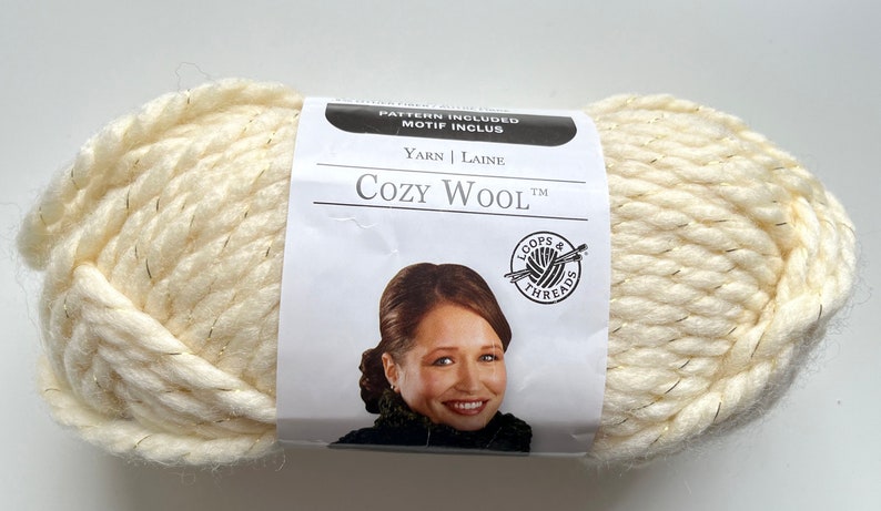 Puede incluir: Ovillo de lana Cozy Wool de color blanco roto con finos hilos met&aacute;licos dorados. La etiqueta dice "Cozy Wool" y muestra la imagen de una mujer. La lana es gruesa y parece suave, para tejer o hacer ganchillo.