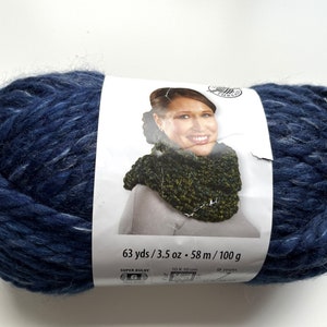 Puede incluir: Una gran madeja de lana azul marino con motas blancas. La etiqueta muestra a una mujer con un cuello tejido. La etiqueta dice "63 yds / 3.5 oz - 58 m / 100 g". La lana es s&uacute;per gruesa y adecuada para tejer o hacer ganchillo.