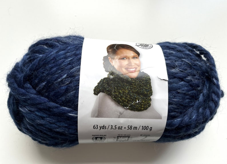 Puede incluir: Una gran madeja de lana azul marino con motas blancas. La etiqueta muestra a una mujer con un cuello tejido. La etiqueta dice "63 yds / 3.5 oz - 58 m / 100 g". La lana es s&uacute;per gruesa y adecuada para tejer o hacer ganchillo.
