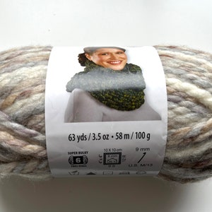 Puede incluir: Ovillo de lana s&uacute;per voluminosa en tonos neutros de beige, gris y blanco. La etiqueta muestra a una mujer con un cuello tejido. La etiqueta indica 58 m, 100 g y 9 mm. Adecuado para agujas de 9 mm.