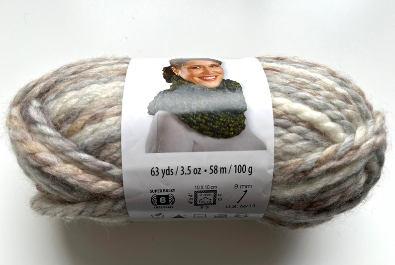 Puede incluir: Ovillo de lana s&uacute;per voluminosa en tonos neutros de beige, gris y blanco. La etiqueta muestra a una mujer con un cuello tejido. La etiqueta indica 58 m, 100 g y 9 mm. Adecuado para agujas de 9 mm.