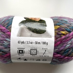Puede incluir: Ovillo de lana s&uacute;per gruesa en tonos morado, azul, amarillo y gris. La etiqueta indica 58 m, 100 g y agujas de 9 mm. La lana es adecuada para agujas del n&uacute;mero 9.