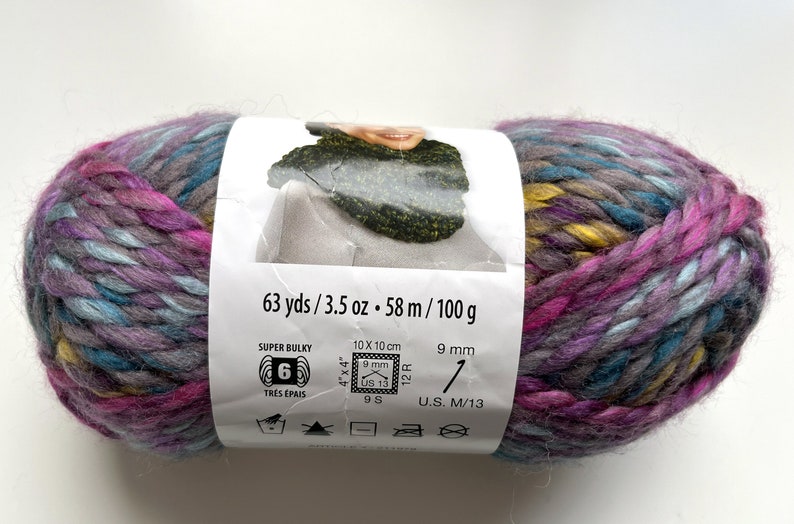 Puede incluir: Ovillo de lana s&uacute;per gruesa en tonos morado, azul, amarillo y gris. La etiqueta indica 58 m, 100 g y agujas de 9 mm. La lana es adecuada para agujas del n&uacute;mero 9.
