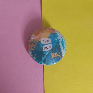 以下が含まれることがあります： 青、オレンジ、白の色合いの抽象的なデザインの丸いボタン。ボタンには、白い長方形の中に「Aro」と「Ace」という文字が入っています。背景はピンクと黄色に分かれています。