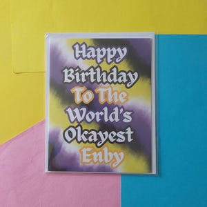 May include: A birthday card with the message "Happy Birthday To The World's Okayest Enby" in white, orange, and purple. The background features a watercolour effect in yellow, purple, and black. The card is in a clear plastic sleeve.