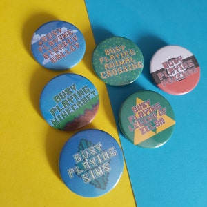 May include: A collection of six round buttons with game-themed designs. Each button features the phrase "Busy Playing" followed by a game title, including "Stardew Valley," "Animal Crossing," "Pokemon," "Minecraft," "Sims," and "Legend of Zelda."