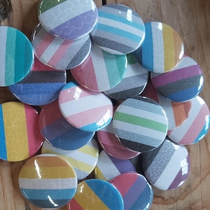 Puede incluir: Una colección de pines de bandera del orgullo coloridos con varios diseños. Los pines son redondos y presentan diferentes combinaciones de colores y patrones.