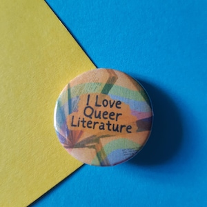 Puede incluir: Un botón colorido con el texto "I Love Queer Literature" sobre un fondo azul y amarillo.