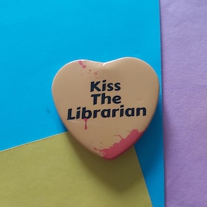 May include: A heart-shaped button with a yellow background and red splatters. The button says "Kiss The Librarian" in black text.