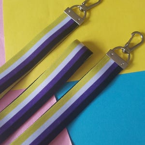 May include: Two non-binary pride flag lanyards with silver metal clasps. The lanyards have horizontal stripes in yellow, white, purple, and black. The background is a mix of yellow, pink, and blue paper.