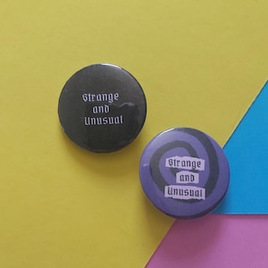 May include: Two round black and purple buttons with the text "Strange and unusual" in white gothic font.
