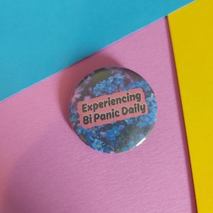 Puede incluir: Un botón redondo con un fondo floral de flores azules y el texto "Experiencing Bi Panic Daily" en un rectángulo rosa. El botón está sobre una superficie rosa con fondos azules y amarillos.