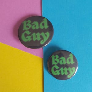 以下が含まれることがあります： 明るい緑色のスタイリッシュな文字で「Bad Guy」と書かれた、2つの丸い濃い灰色のボタン。ボタンはピンク、黄色、青の背景に配置されています。これらのボタンは、どんな服装にも個性を加えるのに最適です。