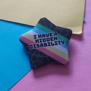 Könnte beinhalten: Ein sternförmiger Button mit dem Text "I HAVE A HIDDEN DISABILITY". Der Button hat ein schwarz-weißes Muster an den Rändern und ein Regenbogenstreifen-Design. Der Hintergrund ist eine Mischung aus Gelb, Blau und Rosa.